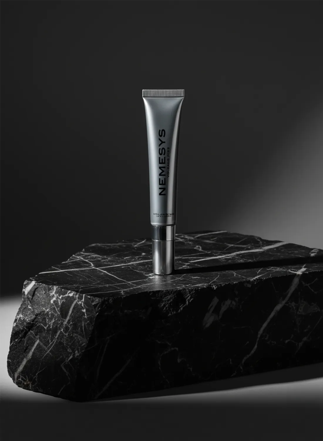 Tube de gloss repulpant NEMESYS posé sur un bloc de marbre noir brut, éclairage minimaliste et ambiance radicale.